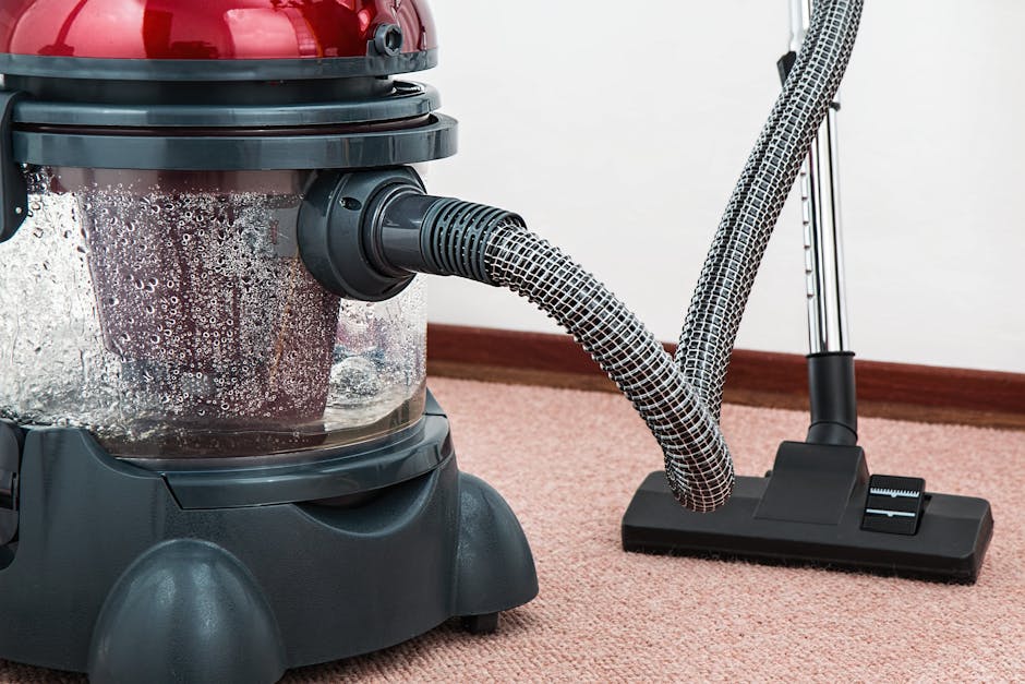 A close-up view of a carpet cleaning vacuum cleaner with a clear dust container filled with dirt and water, connected to a patterned flexible hose. The vacuum is positioned on a pinkish-beige carpet in a room with a white wall and wooden skirting board, reflecting a clean and well-maintained domestic or commercial space. The device appears to be in use or ready for surface cleaning, emphasizing hygiene and deep cleaning processes. There are no visible dust or debris on the carpet, indicating recent or ongoing professional cleaning service by Carpet Cleaners Dulwich, as referenced on the page about surface cleaning and sanitisation in Lordship Lane Dulwich.