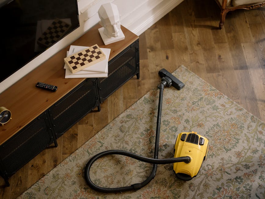 A person dressed in full protective white cleaning suit, including a hood, mask, gloves, and blue shoe covers, is performing deep cleaning on a cream-colored, textured carpet in a well-lit, modern living room. The individual is using a professional carpet cleaning vacuum with a long metal wand to remove dirt and dust from the surface. The room features white walls, wooden furniture, and large potted plants, contributing to a clean and tidy atmosphere. Natural light streams in from a window on the left, emphasizing the freshness and cleanliness of the space. The setting highlights a thorough sanitisation process by Carpet Cleaners Dulwich, demonstrating surface cleaning and maintenance of domestic carpets.