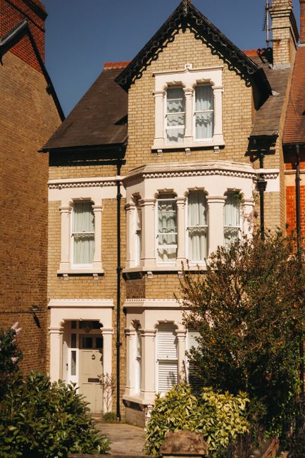 Exterior view of a multi-story residential house with a brick façade and decorative white window frames, situated in East Dulwich SE22. The house features large bay windows on each floor, with some windows having shutters, and a sloped tiled roof with dormer windows. The front garden includes bushes and small trees, with a pathway leading to the white front door. The scene is captured in bright daylight, showcasing the clean and well-maintained appearance of the property. Carpet Cleaners Dulwich, a professional cleaning service, emphasizes interior surface cleaning and deep sanitisation, aligning with end of tenancy cleaning tips for properties like this one, to ensure thorough hygiene and pristine condition.