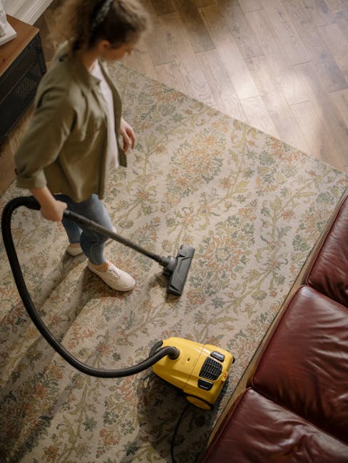 Close-up of a modern vacuum cleaner with a black and gray body, being used to deep clean a patterned area rug with floral motifs in shades of beige, green, and orange, in a well-lit residential living