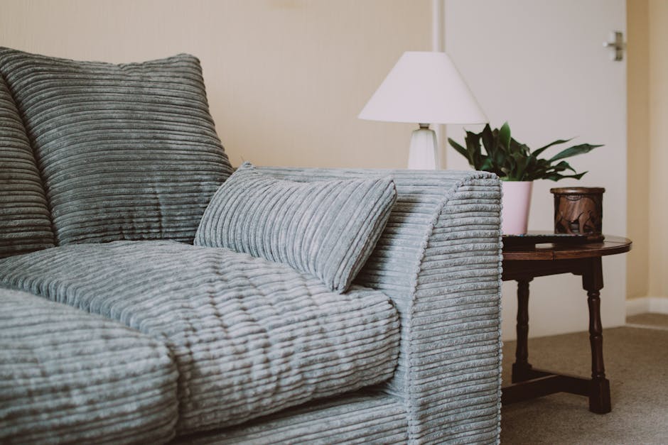A person wearing a dark leather jacket is using a handheld vacuum cleaner to deep clean a decorative pillow with a yellow and white geometric pattern, positioned on a dark fabric sofa in a well-lit in